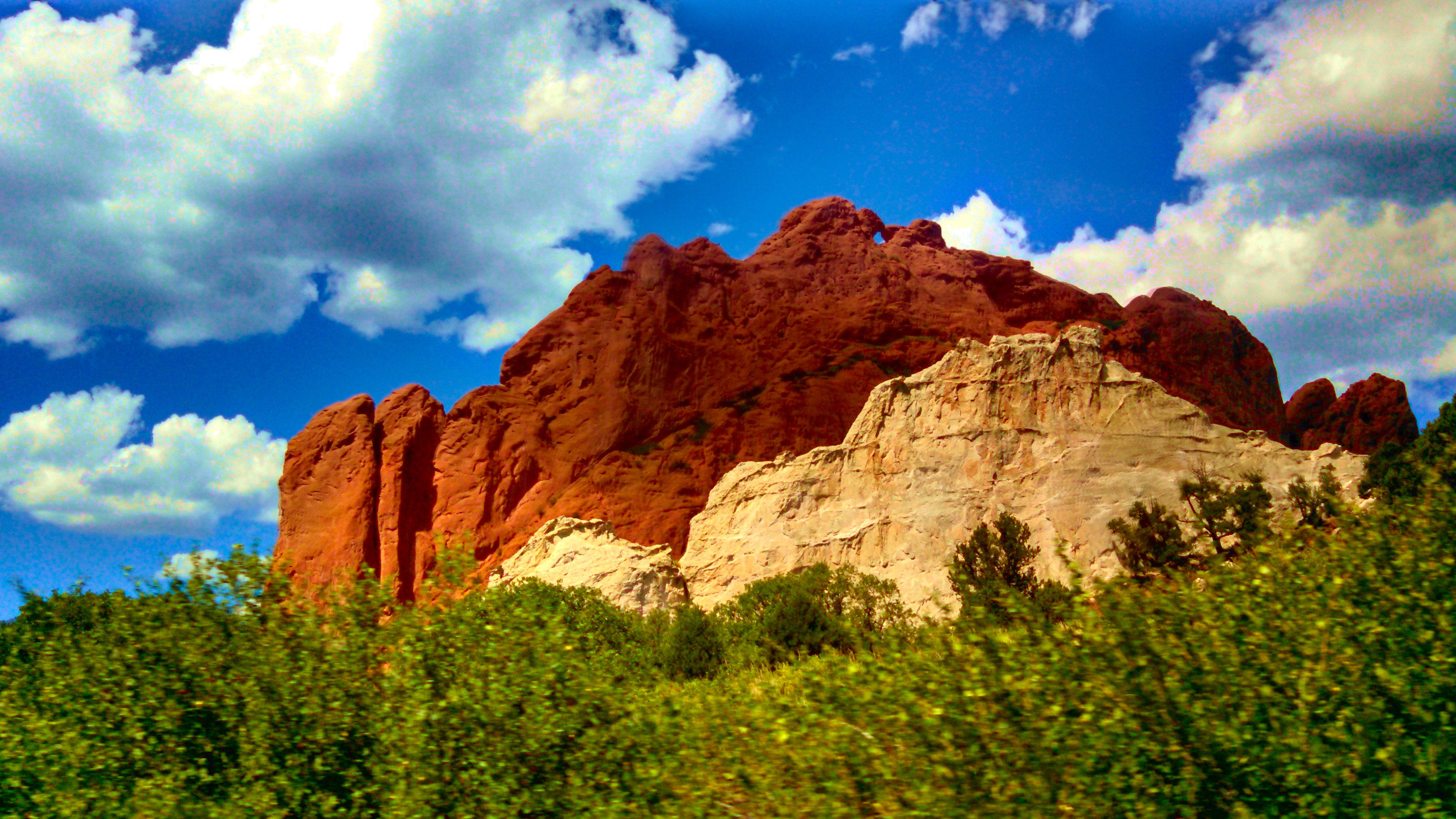 A white rock in front of a red rock, with green foliage in front and a blue sky behind
