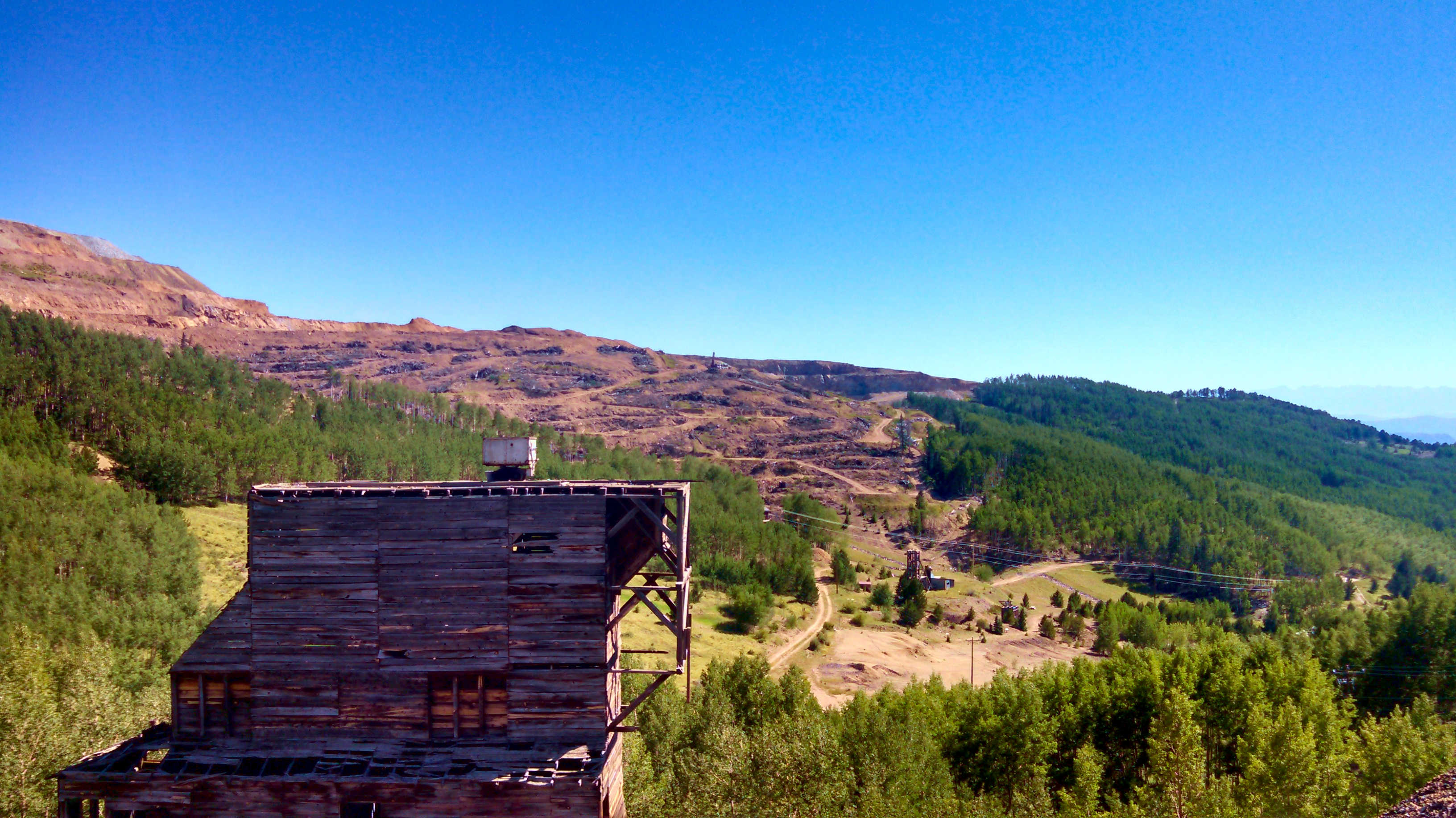 A bare hill is visible in the top left, with green trees on all the other ground. There are the ruins of an old wooden building in the bottom left
